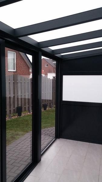 Die Terrasse ist mit einem schwarzen Rahmen, einer Glastür und einem Fenster versehen. Der Innenraum ist mit hellen Fliesen ausgelegt, der Außenbereich besteht aus einem Ziegelweg, Rasen und einem Zaun.