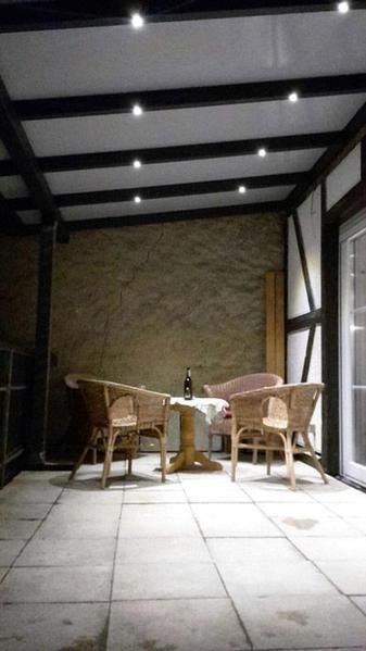 Terrasse mit Rattanmöbeln, einem kleinen Tisch, einer Steinmauer und Deckenbeleuchtung.