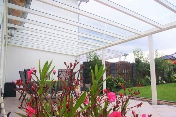 Überdachte Terrasse mit transparentem Dach und weißem Rahmen, mit Blick auf einen grünen Hof und Garten.
