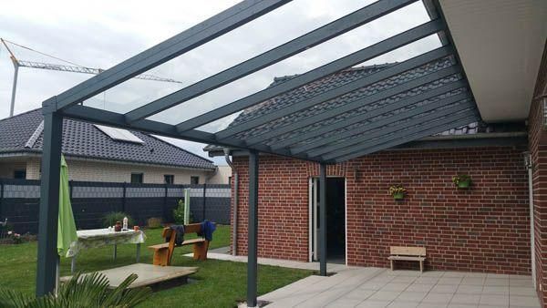 Graue Pergola mit Glasdach, die an ein Backsteinhaus angebaut ist, über einer Terrasse und einem grasbewachsenen Hinterhof.