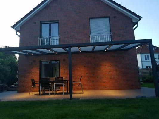 Terrasse mit Pergola, Sitzgelegenheiten und Beleuchtung, an ein Backsteinhaus angebaut. Nachtszene.