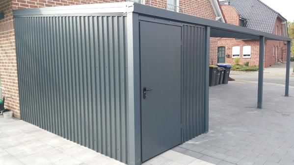 Grauer Metallschuppen mit Tür und angebautem Carport auf einer gepflasterten Fläche neben einem Backsteingebäude.