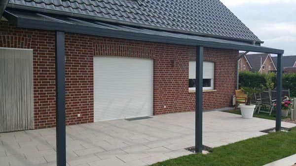 Eine graue Metall-Terrassenüberdachung ist an ein rotes Backsteinhaus mit weißem Garagentor angebaut und befindet sich auf einer gefliesten Terrasse.