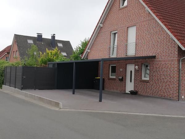 Carport an einem roten Backsteinhaus; dunkelgraues Gebäude mit gepflasterter Zufahrt.