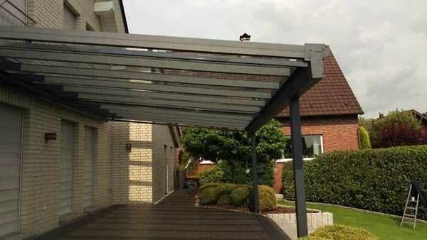 Überdachte Terrasse mit Glasdach, die an ein helles Backsteinhaus und einen grünen Garten angrenzt.