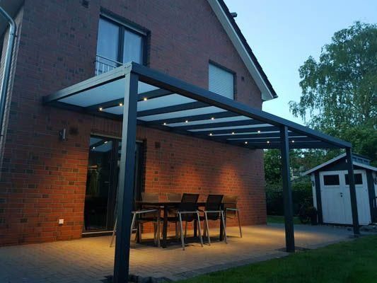 Terrasse mit schwarzer Pergola, Beleuchtung, Tisch, Stühlen und einem Backsteinhaus im Hintergrund.