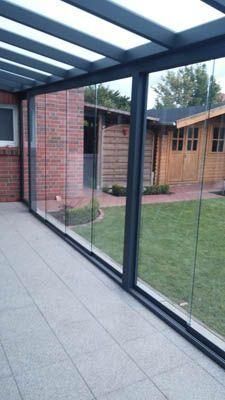 Verglaste Terrasse mit schwarzem Rahmen, Fliesenboden, Backsteinmauer und einem Holzschuppen im Hintergrund.