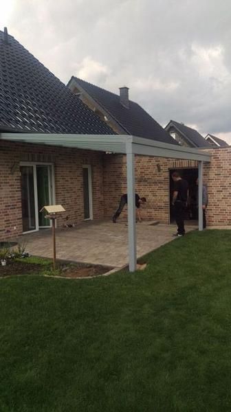 Eine Terrasse mit einer weißen Pergola, die an ein Backsteinhaus angebaut ist. Grüner Rasen und ein bewölkter Himmel.