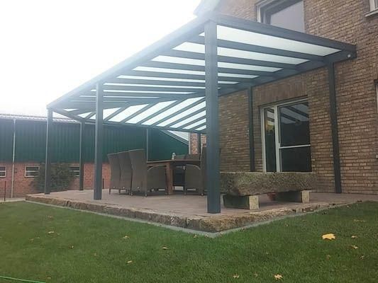 Terrasse mit schwarz umrahmter Pergola, Backsteinhaus, Gartenmöbeln und Rasenfläche.