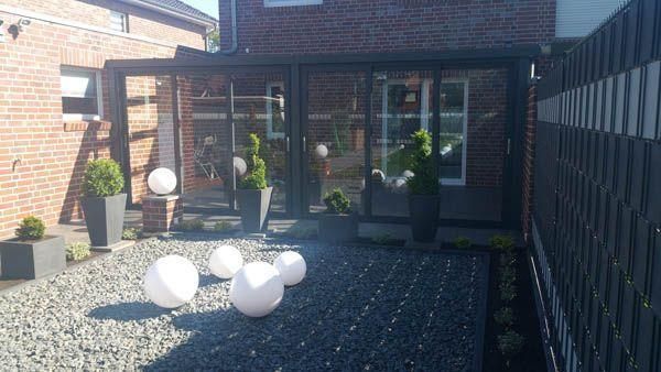 Terrasse im Freien mit Glaswänden, Ziegel- und dunkelgrauen Rahmen, leuchtenden Kugeln, Zierpflanzen und dunklem Stein.