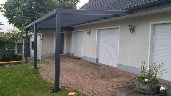Eine Pergola, die an ein helles Haus über einer Ziegelterrasse mit heruntergelassenen Garagentoren angebaut ist.