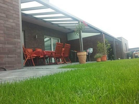 Grüner Rasen vor einem Backsteinhaus mit Terrasse, Tisch, Stühlen und Topfpflanzen unter einem Vordach.