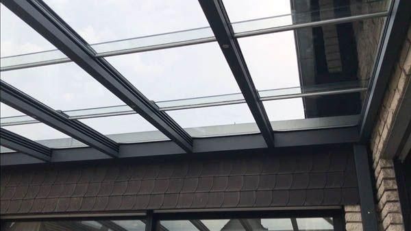 Ein von dunklen Metallträgern getragenes Glasdach vor einer Backsteinmauer und bedecktem Himmel.