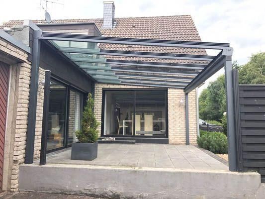 Eine moderne graue Pergola mit Glasdach überspannt eine Terrasse vor einem Backsteinhaus.
