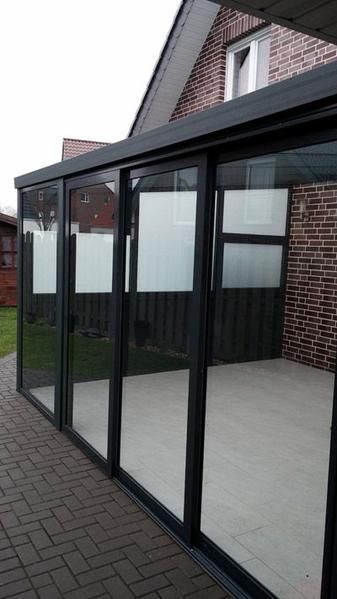 Eine verglaste, mit schwarzen Rahmen versehene Terrasse ist an ein Backsteingebäude angebaut. Helle Bodenbeläge und ein bewölkter Himmel sind zu sehen.