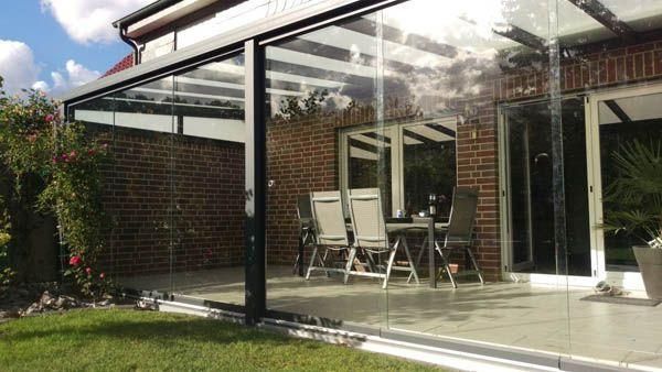 Verglaste Terrasse mit schwarzem Rahmen, Backsteingebäude im Hintergrund, Tisch und Stühle im Inneren.