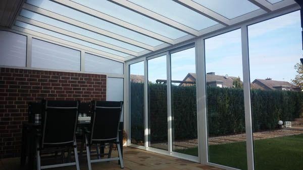 Wintergarten mit Glaswänden und -dach, neben einer Backsteinmauer und einer Essgruppe im Freien, mit Blick auf einen grünen Garten.