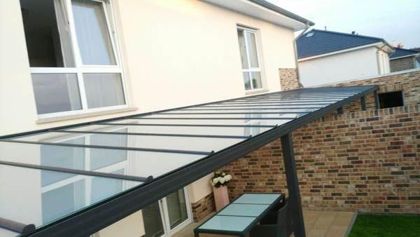 Eine überdachte Terrasse mit Glasdach, Tisch und Stühlen, die an eine Backsteinmauer und ein cremefarbenes Haus angrenzt.