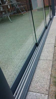 Dreigleisige Glasschiebetüren mit grauem Rahmen, neben einer Steinterrasse.