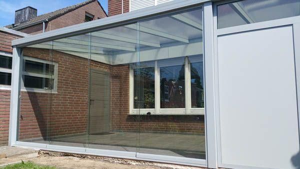 Verglaster Wintergarten an einem Backsteinhaus. Grauer Rahmen, Klarglas und eine weiße Seitenwand.