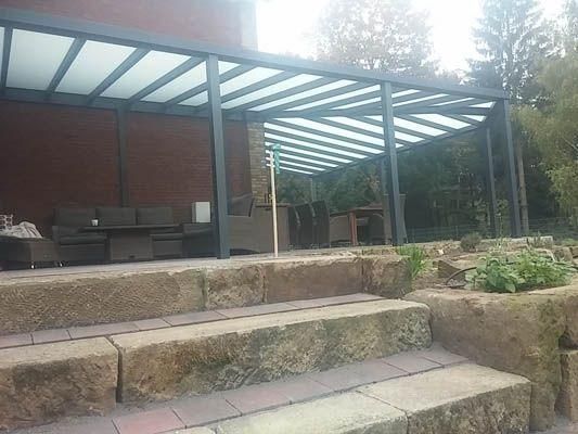 Steinstufen führen zu einer Terrasse mit einem Pergola-Dach aus Metallrahmen. Darunter stehen Gartenmöbel.