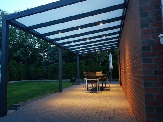 Terrasse mit dunklem Rahmen, weißem, lichtdurchlässigem Dach, eingebauter Beleuchtung, Backsteinmauer sowie Tisch und Stühlen.