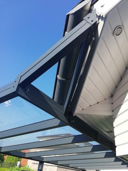 Ein Vordach aus Glas und Metall, das an einem Gebäudedach befestigt ist, vor blauem Himmel.