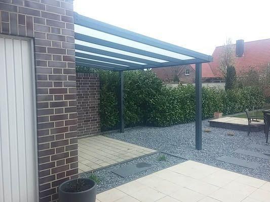 Eine überdachte Terrasse, die an ein Backsteingebäude angebaut ist. Graues Gerüst, lichtdurchlässiges Dach, Kiesboden und üppige grüne Büsche.