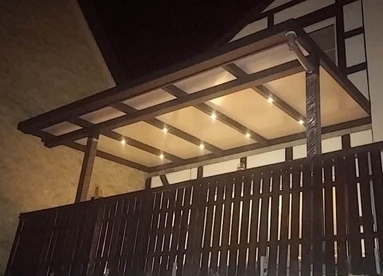 Eine hölzerne Pergola mit Lichterketten über einem dunkelbraunen Zaun. Nachtszene.