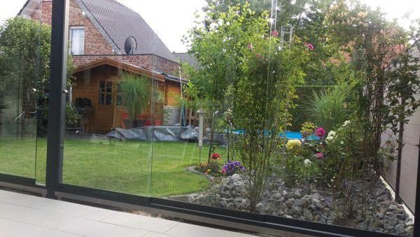 Ein Garten, betrachtet durch Glasscheiben. Grüner Rasen, Bäume, Blumen und ein Holzschuppen sind zu sehen.