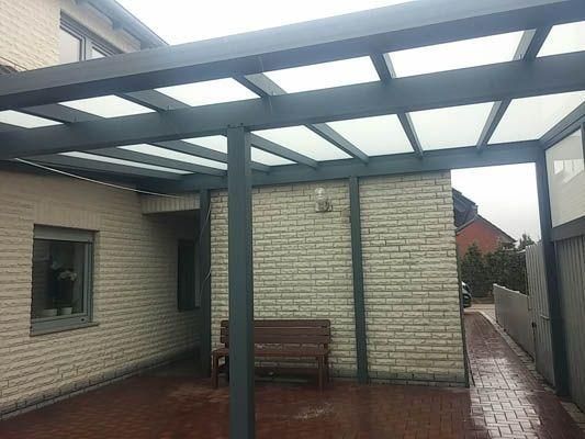 Eine grau umrahmte Pergola mit transparentem Dach überspannt eine Terrasse neben einem Backsteinhaus. Darunter steht eine Bank.