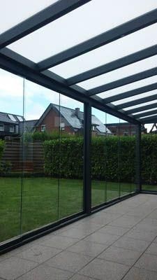 Verglaste Terrassenüberdachung mit grauem Rahmen und Dach, die auf einen Hinterhof mit grünem Rasen, Hecken und Häusern blickt.