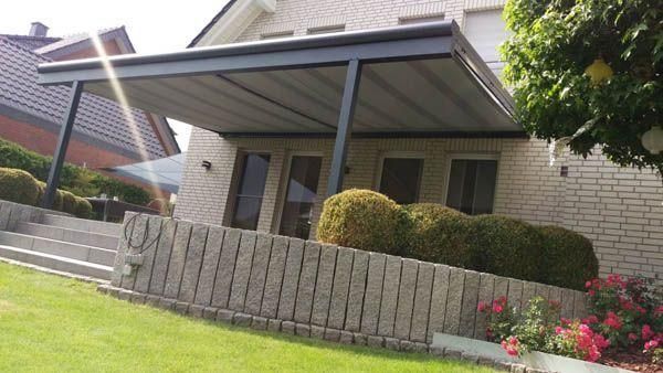 Eine moderne Pergola spendet Schatten auf einer Terrasse vor einem Backsteinhaus. Im Vordergrund sind Stufen und eine Stützmauer zu sehen.