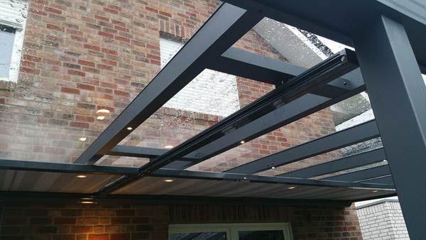 Eine Terrasse mit dunkelgrauem Metallrahmen, transparenten Dachplatten und eingebauter Beleuchtung vor einer Backsteinmauer.