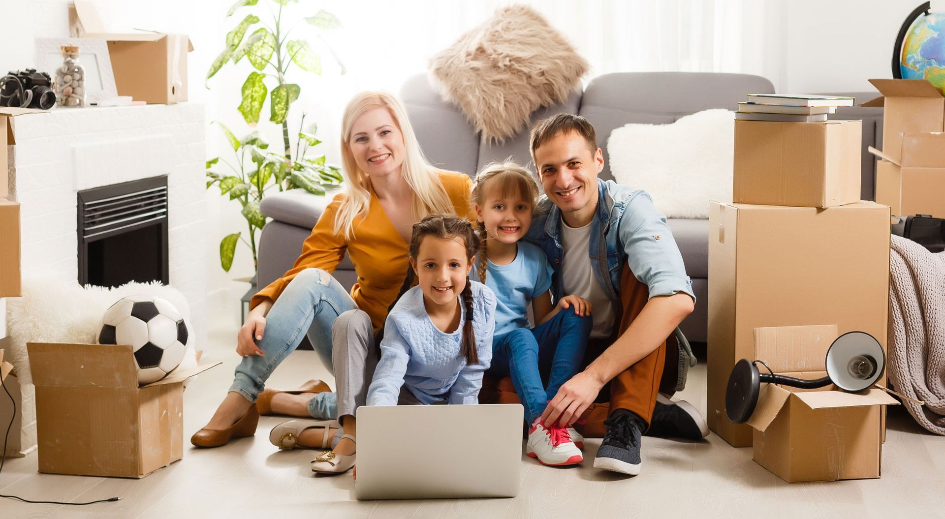 Eine vierköpfige Familie lächelt und sitzt auf dem Boden, umgeben von Umzugskartons, einem Laptop und einem Fußball.