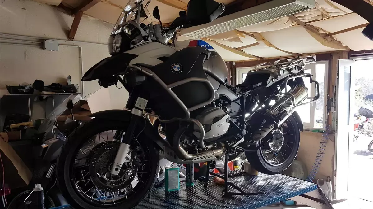 Deux motos noires garées dans un garage encombré, dont une sur une plateforme métallique près d’une porte lumineuse.