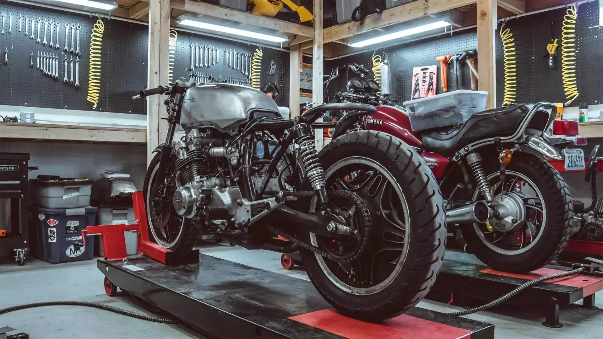 Deux motos personnalisées sur des ponts élévateurs dans un atelier de garage, avec des outils et des étagères en arrière-plan
