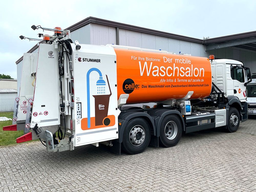 Vor einem Gebäude parkt ein weiß - orangefarbener Müllwagen.