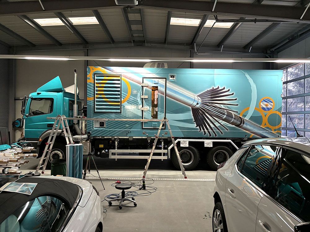 Ein blauer Lastwagen steht in einer Garage neben einem weißen Auto.