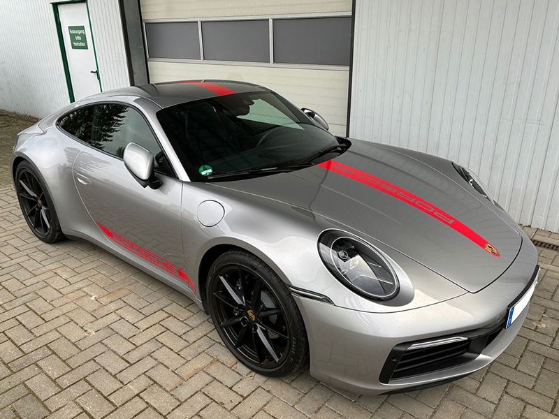 Ein silberner Porsche 911 mit einem roten Streifen auf der Motorhaube steht auf einem gepflasterten Gehweg.