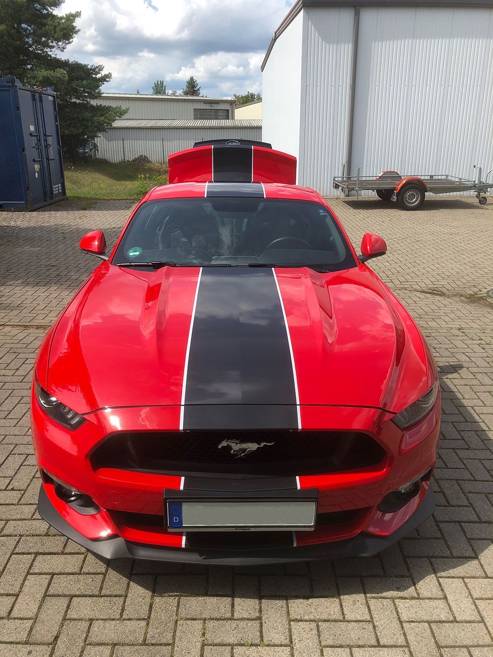 Ein roter Mustang mit schwarzen und weißen Streifen auf der Motorhaube ist auf einer gepflasterten Auffahrt geparkt.