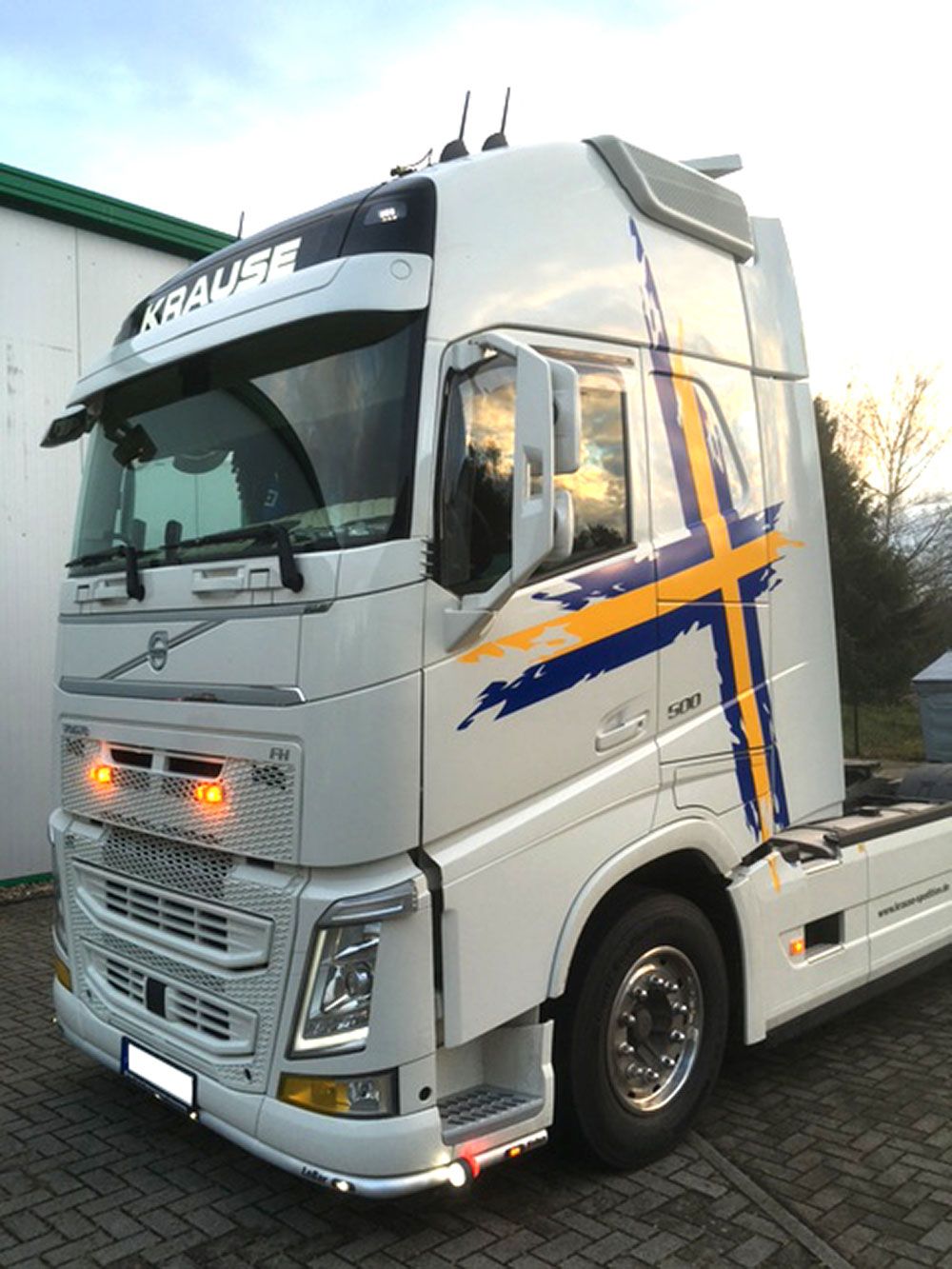 Ein weißer Sattelschlepper mit einem blau-gelben Kreuz an der Seite steht vor einem Gebäude.