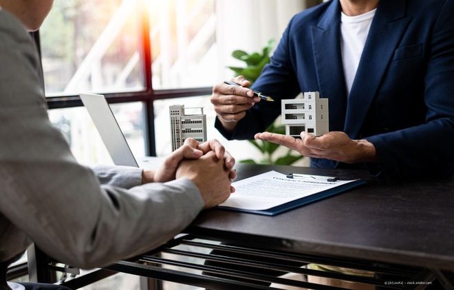 Zwei Personen unterhalten sich an einem Tisch über Immobilienmodelle; eine von ihnen hält einen Stift und Dokumente in der Nähe.