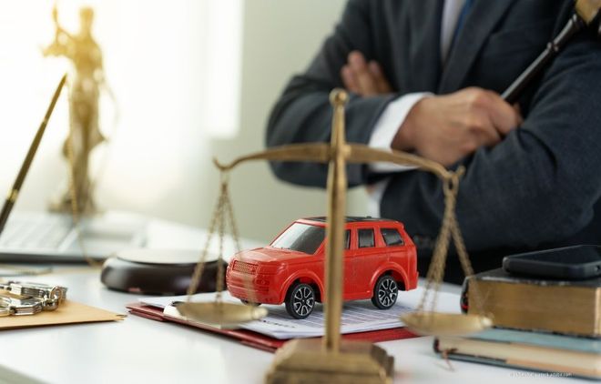 Ein rotes Spielzeug-SUV steht auf einem Schreibtisch neben einer Justitia-Waage aus Messing; im Hintergrund ist ein Anwalt zu sehen.