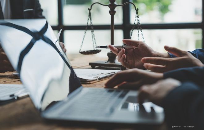 Ein Anwalt prüft Dokumente an einem Schreibtisch mit einem Laptop; im Hintergrund ist eine Justitiawaage zu sehen.