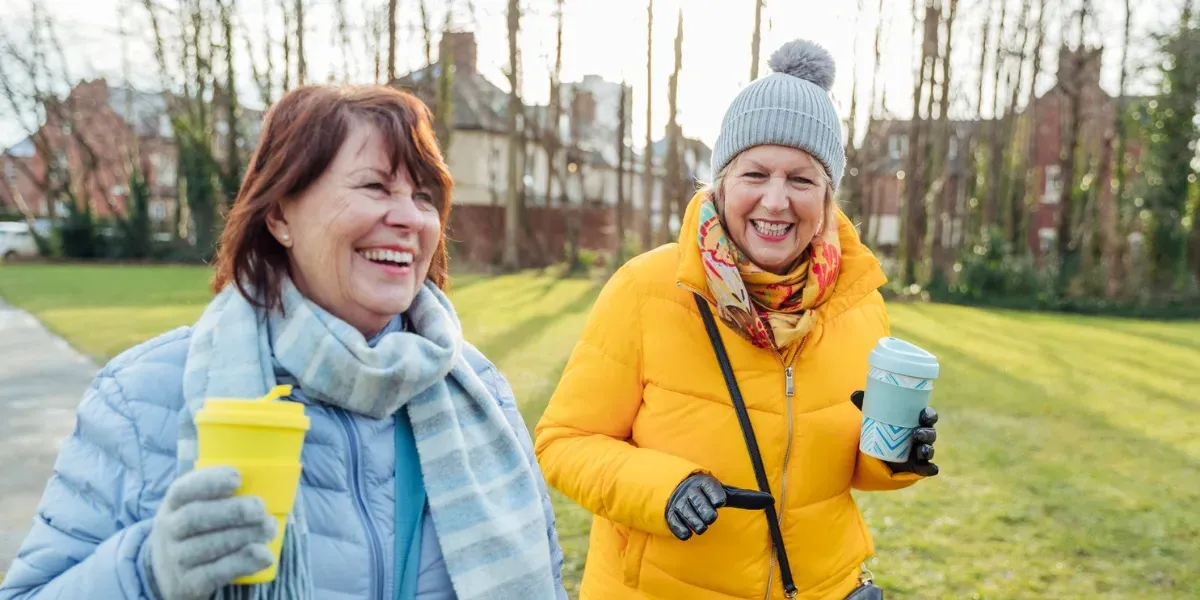Two people walking outside, smiling and holding drinks. One wears a yellow coat and hat.