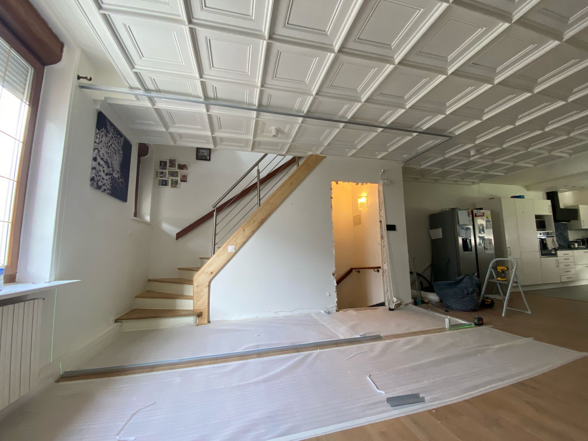Pièce en rénovation avec escalier, revêtement de sol recouvert de ruban adhésif, porte ouverte, échelle et lumière.