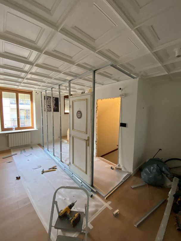 Pièce en rénovation avec charpente apparente, plafond lambrissé blanc, éclairage par fenêtre et outils de construction au sol.
