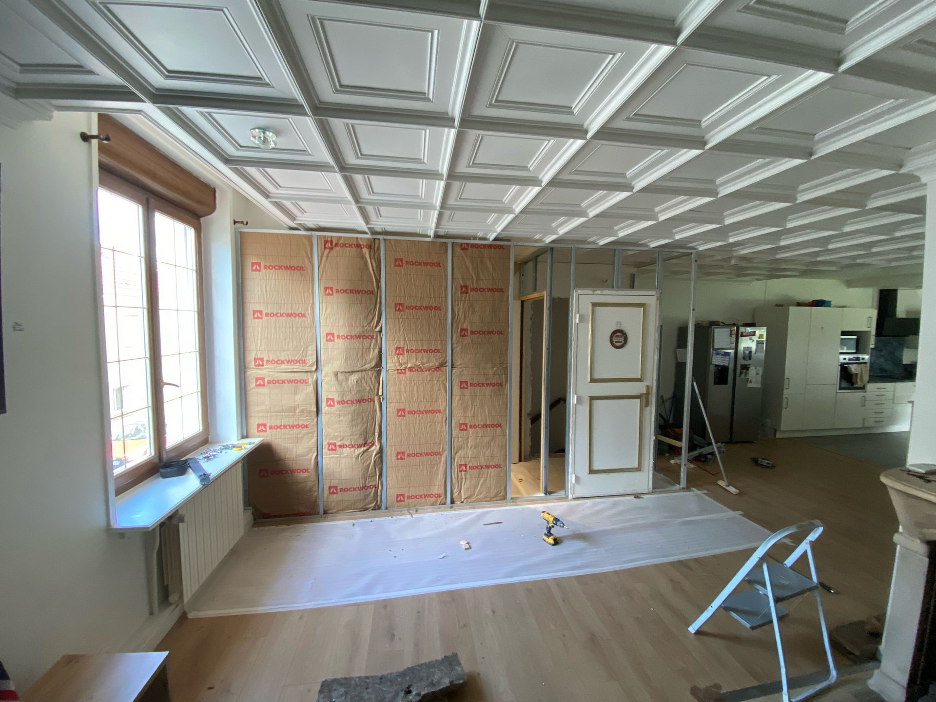 Pièce vide en cours de rénovation avec plafond à caissons, isolation apparente, fenêtres et outils éparpillés.