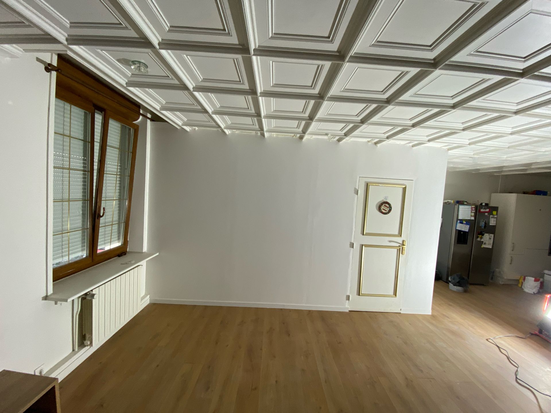 Pièce vide avec parquet, plafond orné, grandes fenêtres et une porte blanche sur le mur du fond.